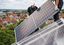 Zwei Personen installieren ein Solarpanel auf einem Dach. Im Vordergrund ist das Solarpanel schräg zu sehen, während im Hintergrund eine Person in dunkler Arbeitskleidung mit weißem Helm und eine weitere Person das Panel halten und positionieren. Im Hintergrund erstreckt sich eine Stadt mit roten Ziegeldächern, grünen Bäumen und bewölktem Himmel.