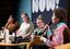 Das Bild zeigt eine Podiumsdiskussion mit vier Frauen, die auf Stühlen sitzen und Mikrofone in der Hand halten. Alle sind in ein Gespräch vertieft, und eine Person spricht gerade ins Mikrofon. Im Hintergrund ist ein großes Schild mit dem Schriftzug „NOW!“ zu sehen. Die Teilnehmer:innen haben unterschiedliche, farbenfrohe Kleidung an, und es befinden sich Wasserflaschen, Bücher und Notizblöcke auf den kleinen Tischen vor ihnen. Die Atmosphäre wirkt konzentriert und professionell, als würden wichtige Themen besprochen.