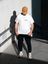 Ein farbiges Foto einer Person mit brauner Kappe, weißem T-Shirt und schwarzer Hose. Auf dem T-Shirt stehen neben einem orangefarbenen Zeichen die Worte „Coffee Bytes“. Die Person steht im Sonnenlicht vor einer Betonwand.