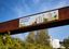 Großformatiges Banner des Volksparks Potsdam an einer rostfarbenen Brücke, gestaltet mit farbenfrohen Illustrationen von Pflanzen, spielenden Menschen und Schaukeln vor blauem Himmel.